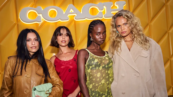 Four women posing in front of a yellow background with the illuminated Coach logo, wearing fashionable outfits.